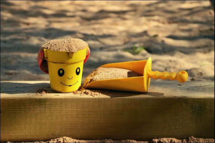 Close up of a yellow toy bucket and shovel filled with sand next to a sandbox