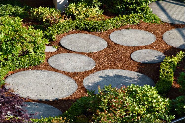 Small space with red soil and grey round pavers placed randomly on it, surrounded by green bushes