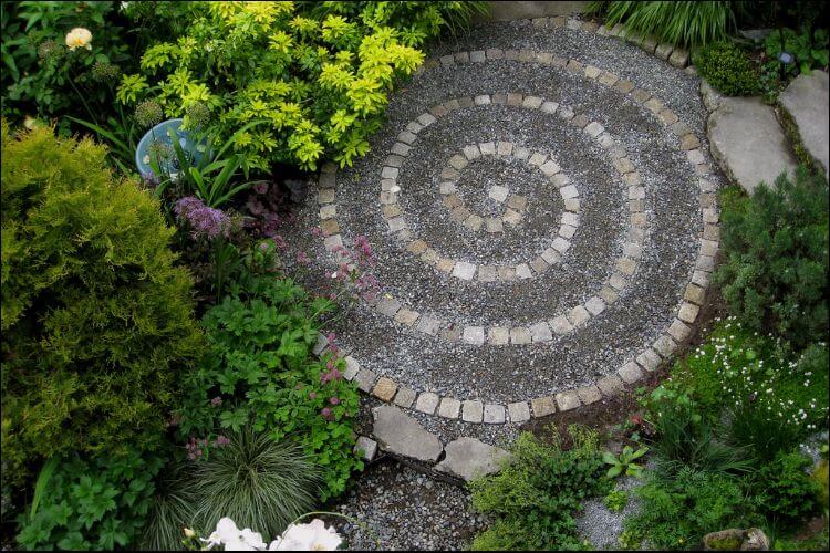 A round focal point with a spiral pattern made of gray and white pavers, surrounded by green bushes and flowers