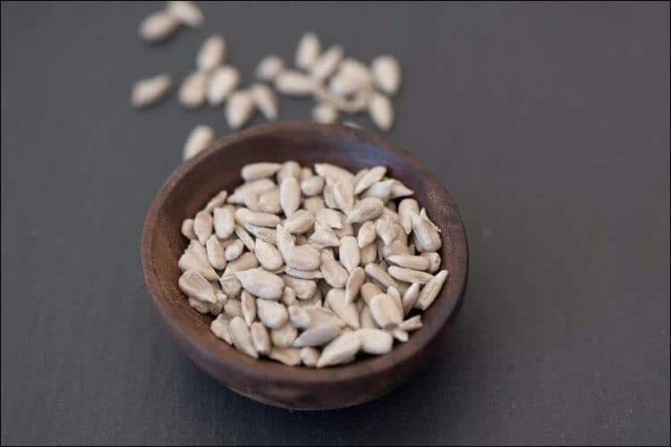 Close up of peeled sunflower seeds in a small dark wooden bowl, with other seeds visible in the blurry background
