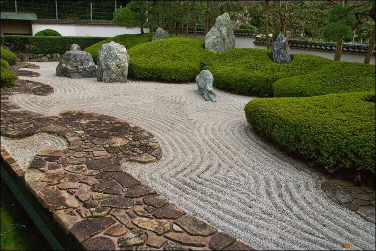 Winding pathway made of colored pavers, placed next to a larger gravel pathway, surrounded by green thick bushes with big boulders placed here and there