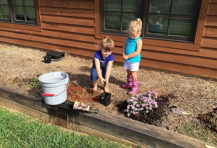 two girls planting flowers in earth outside a house
